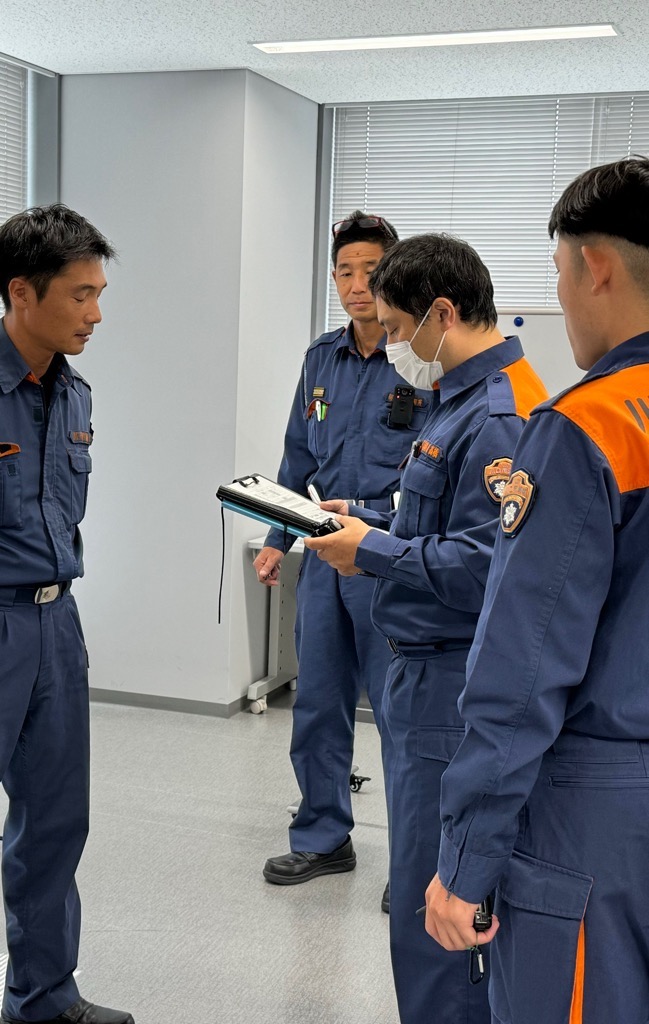 4人の消防隊員が立って、一人が関係者役の者に聞き取りをしている写真