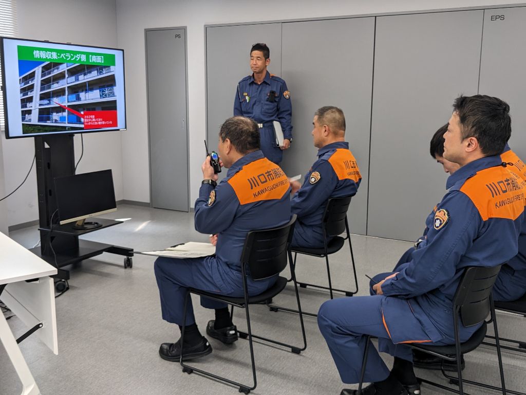 消防車内を想定して椅子に座った4人の消防隊員。そのうちの一人は隊長として無線機で報告し、みんなでモニター画面を見ている写真。