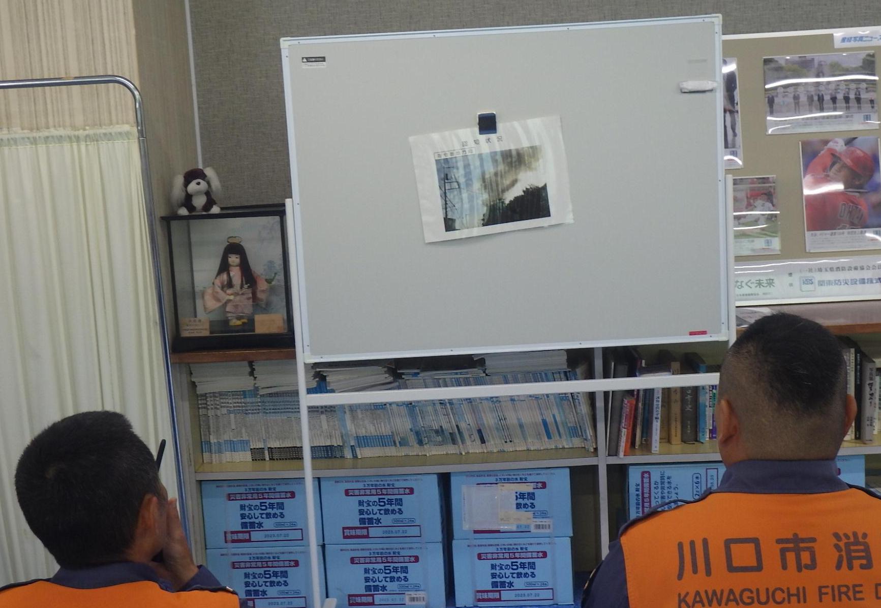 現場に向かう途中の報告。ホワイトボードに張り出された画像を見て、煙の色、煙が流れている方向など警防本部に報告します。