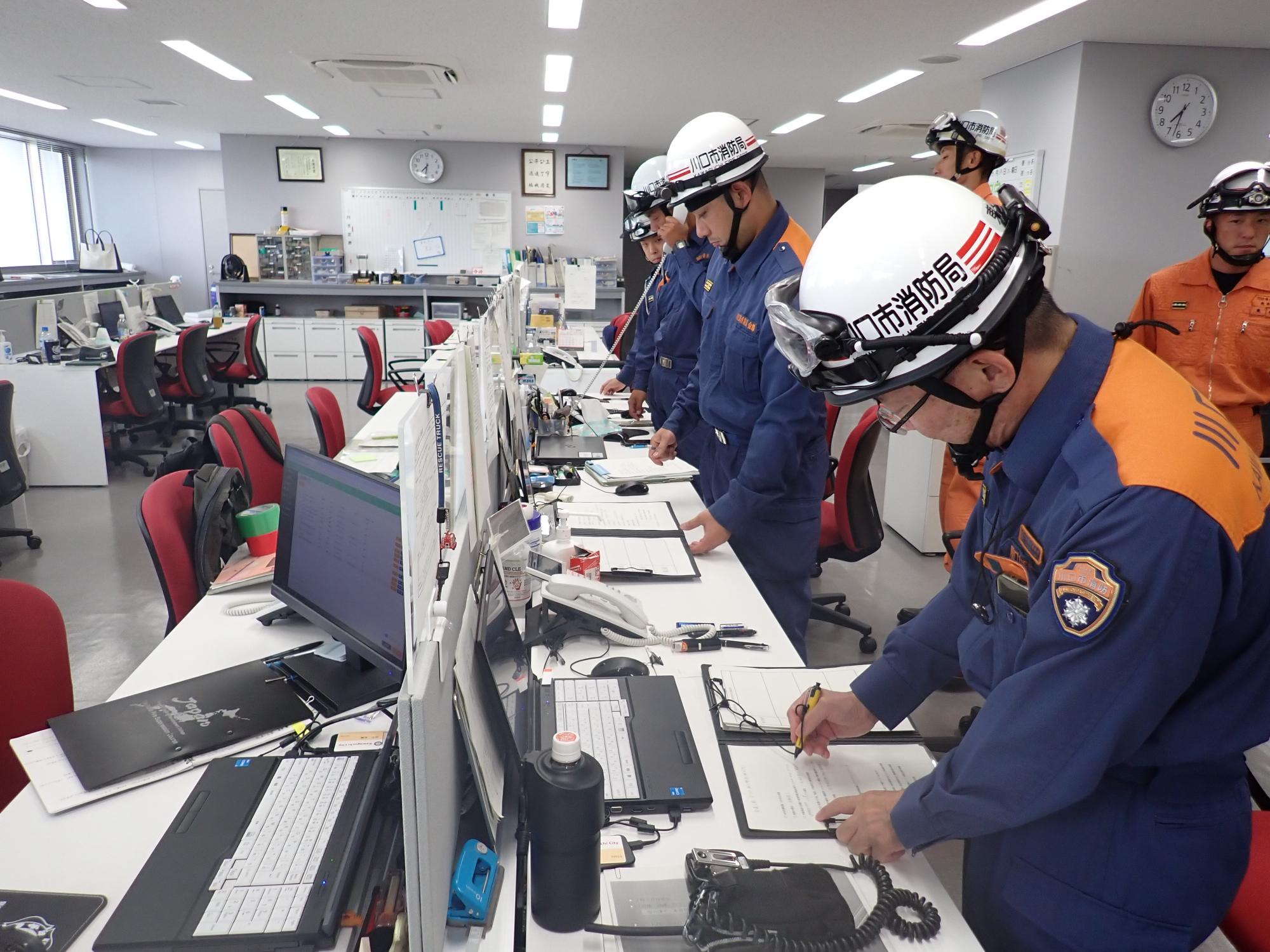 東消防署の事務所内で、消防隊がヘルメットをかぶりメモを取っている写真