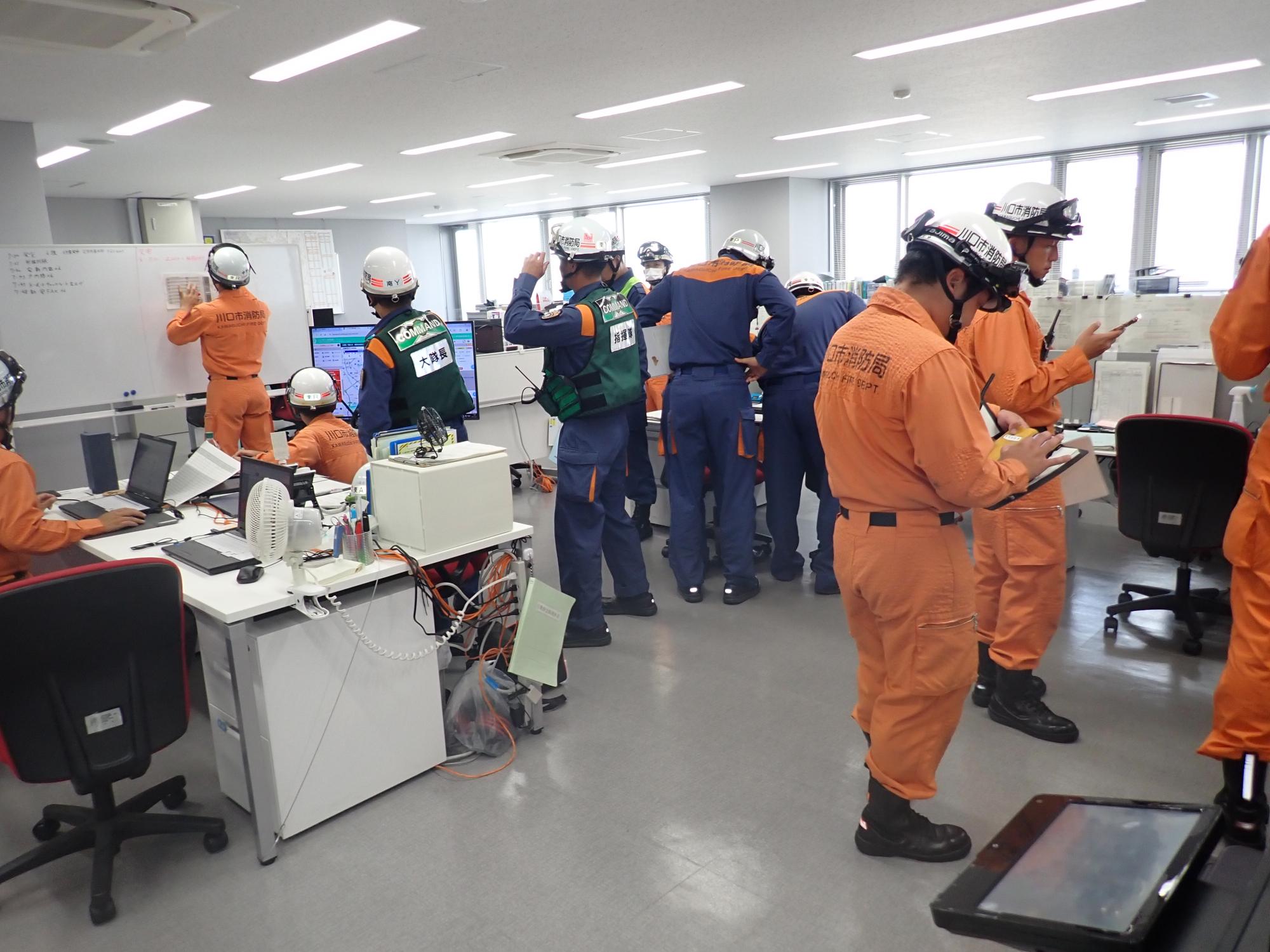 東消防署の事務所内で、10人以上の消防隊員・救助隊員がメモをとるなどし、情報収集をしている写真