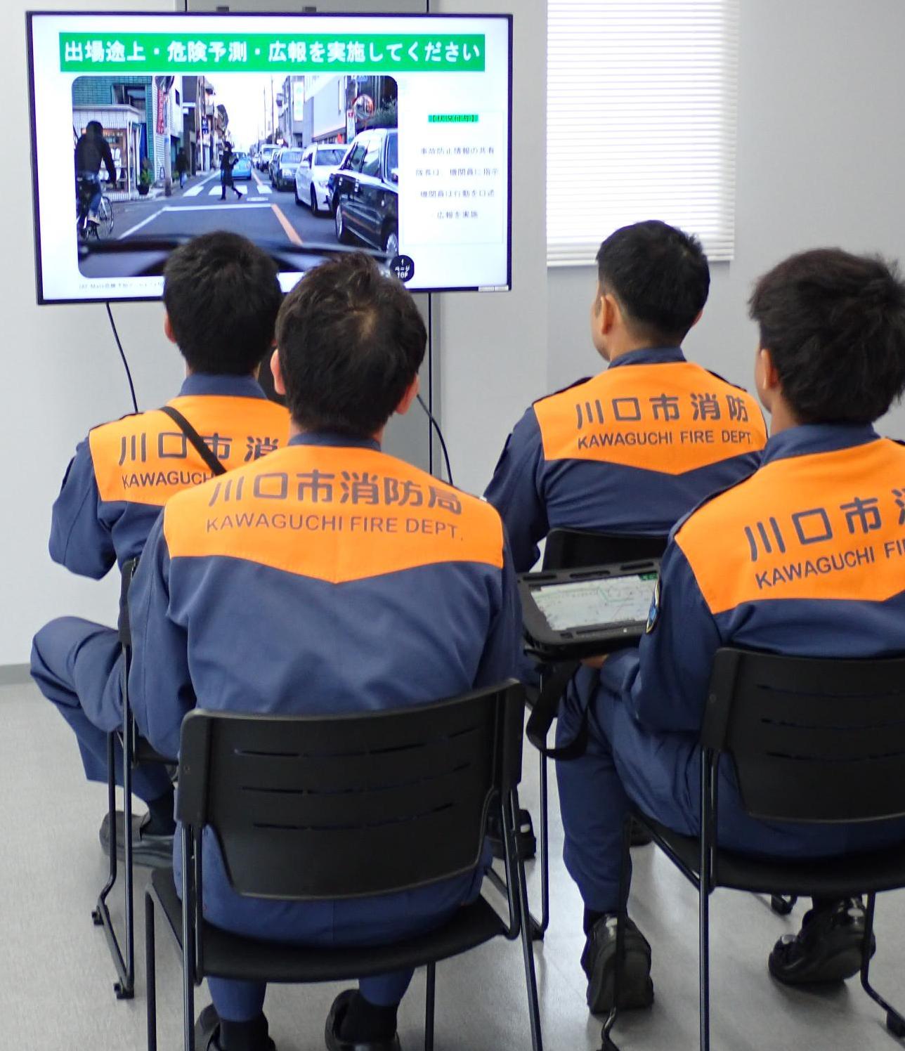 車の配置と同じに置いた椅子に座った4人の隊員が、表示された道路の場面を見ている写真